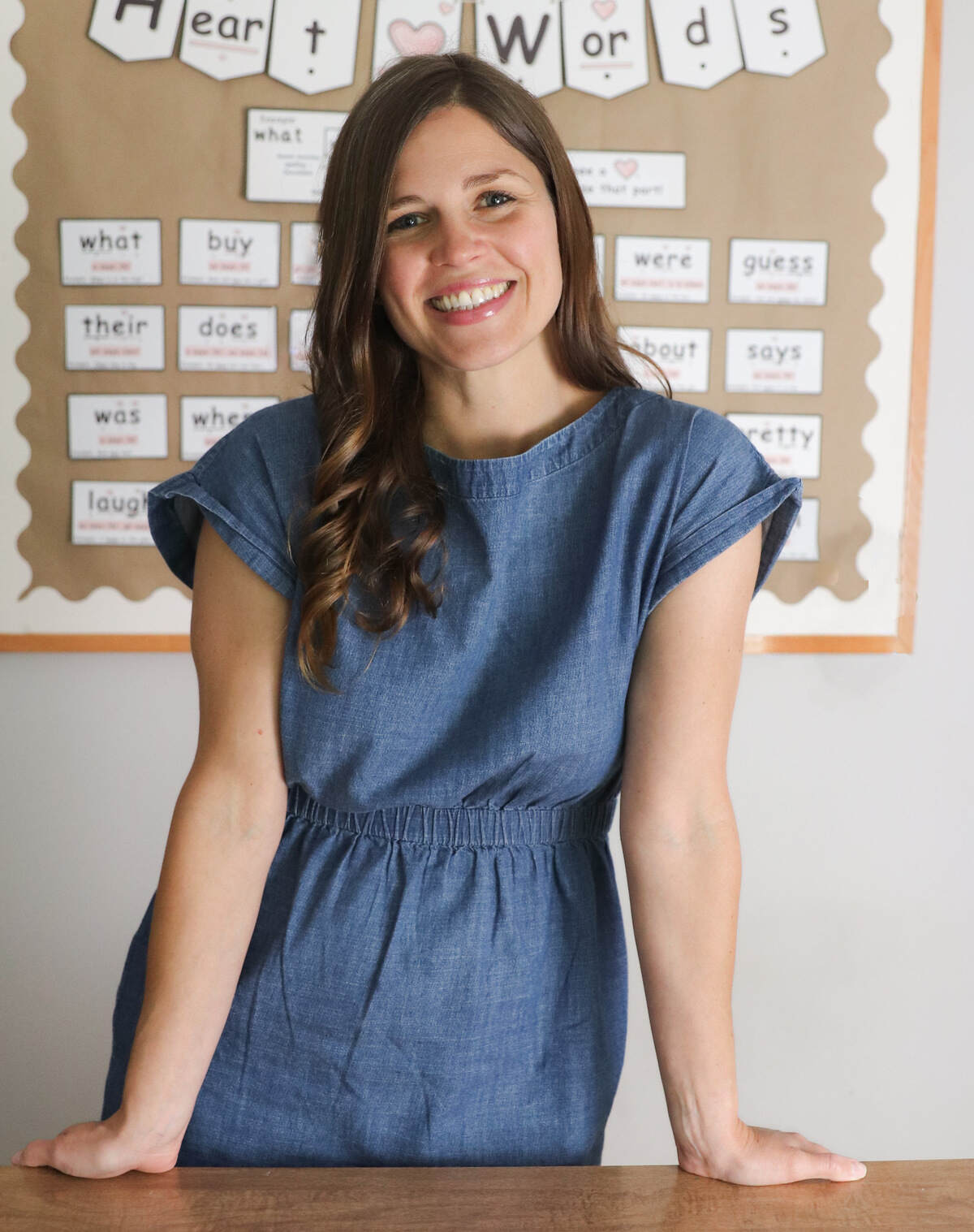 A smiling brown haired woman in a denim dress in a classroom.
